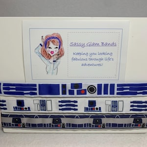 May include: Three rolls of white and blue patterned ribbon with a Star Wars R2-D2 design. The ribbon is displayed on a white board with a card that reads "Sassy Glam Bands keeping you looking fabulous through life's adventures!"