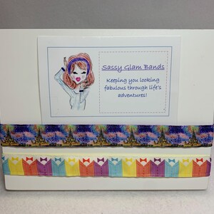 May include: A white card with a cartoon illustration of a woman with brown hair wearing a blue headband and a blue shirt. The card has the text "Sassy Glam Bands" and "Keeping you looking fabulous through life's adventures!"  The card is displayed on a white stand with two colorful patterned ribbons.