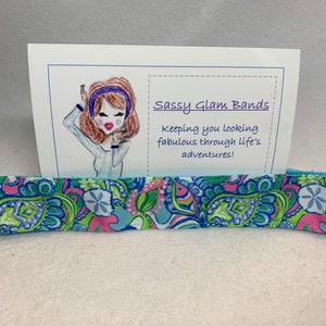 May include: A patterned headband with a blue, green, and pink design. The headband is decorated with a paisley and floral pattern. The text on the card reads "Sassy Glam Bands" and "Keeping you looking fabulous through life's adventures!"