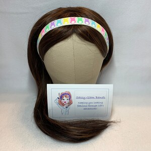 Puede incluir: Una diadema blanca con siluetas de conejos pastel de colores. La diadema est&aacute; en un maniqu&iacute; de peluca marr&oacute;n. El texto en la tarjeta dice "Sassy Glam Bands, Keeping you looking fabulous through life's adventures!"
