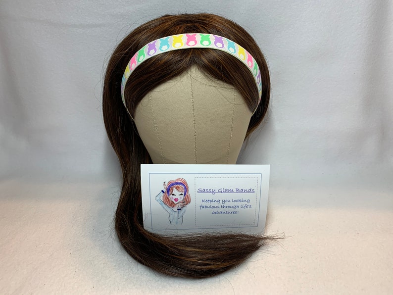 Puede incluir: Una diadema blanca con siluetas de conejos pastel de colores. La diadema est&aacute; en un maniqu&iacute; de peluca marr&oacute;n. El texto en la tarjeta dice "Sassy Glam Bands, Keeping you looking fabulous through life's adventures!"