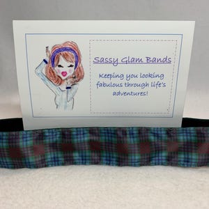 Może przedstawiać: Opaska do włosów w niebiesko-zielono-bordową kratę z czarną gumką. Opaska jest prezentowana na białym tle z kartą, na której widnieje napis "Sassy Glam Bands" i "Keeping you looking fabulous through life's adventures!"