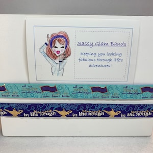 Può includere: Un pannello di visualizzazione bianco con un biglietto che recita "Sassy Glam Bands Keeping you looking fabulous through life's adventures!" Il biglietto presenta un'illustrazione a fumetti di una donna con i capelli rossi che indossa una fascia blu. Due nastri blu e dorati con il testo "Diamond in the rough" e "Tell me now, when did you your heart decide" sono esposti sul pannello.