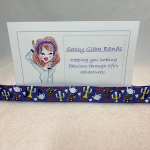 Könnte beinhalten: Ein blaues Stirnband mit einem sich wiederholenden Muster aus weißen Teetassen, braunen Kerzenständern und braunen Figuren. Das Stirnband wird vor einer Karte mit dem Text "Sassy Glam Bands" präsentiert. Die Karte hat auch eine Zeichnung einer Frau.