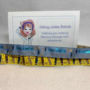 Könnte beinhalten: Zwei gelbe und blaue Haargummis mit dem Text "I want adventure" darauf gedruckt. Die Haargummis liegen auf einer weißen Oberfläche mit einer Karte, auf der "Sassy Glam Bands" und "Keeping you looking fabulous through life's adventures!" steht.