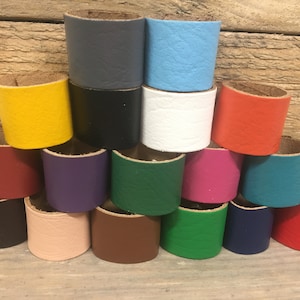 May include: A selection of colourful leather napkin rings in shades like yellow, black, white, red, and blue. The rings are stacked, displaying their cylindrical form and textured surface. The background is a light-coloured wooden surface.