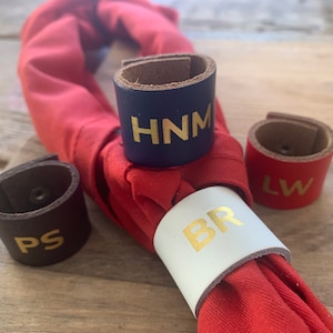 May include: A selection of leather napkin rings in navy, red, white, and brown. Each ring has gold lettering with initials. The rings are around a red napkin, perfect for a dining table setting.