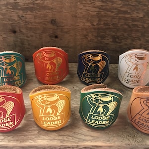 May include: Eight Lodge Leader patches in various colours, including teal, orange, navy, and white. Each patch features a cartoon beaver and the words "Lodge Leader" in gold lettering. The patches are displayed against a wooden background.