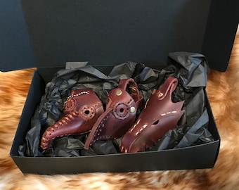 May include: Three brown leather plague doctor masks in a black box with black tissue paper. The masks have a long beak and are decorated with stitching and metal accents.