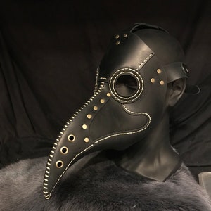 May include: A black leather plague doctor mask with a long beak, white stitching, and gold-toned rivets. The mask is on a mannequin head.