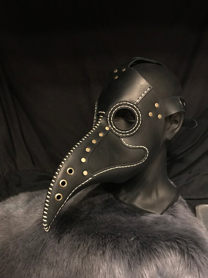 May include: A black leather plague doctor mask with a long beak, white stitching, and gold-toned rivets. The mask is on a mannequin head.