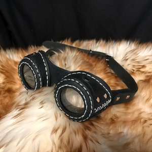 Puede incluir: Gafas steampunk de cuero negro con costuras blancas y detalles dorados. Las gafas tienen lentes redondas y una correa.