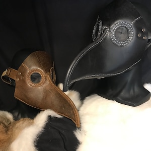 May include: Two leather plague doctor masks, one brown and one black, with stitched eye holes and a long beak. The masks are displayed on a white fur surface.
