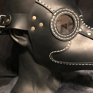 May include: Black leather plague doctor mask with a white stitched design and a clear plastic eye piece. The mask has a strap for wearing.