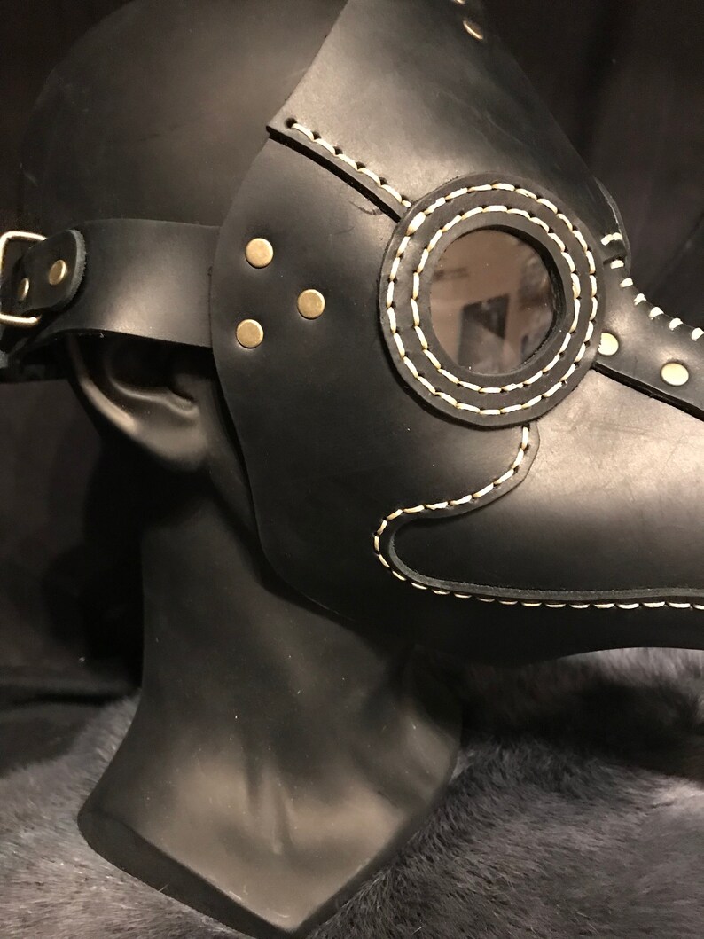 May include: Black leather plague doctor mask with a white stitched design and a clear plastic eye piece. The mask has a strap for wearing.