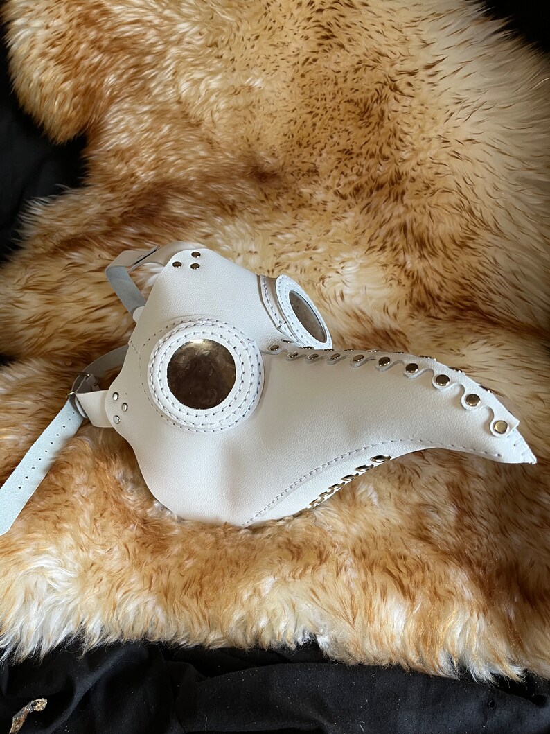 May include: White leather plague doctor mask with two round eye pieces and a long beak. The mask has metal rivets and stitching details.