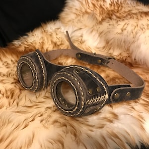 Puede incluir: Gafas steampunk de cuero marrón con lentes redondos y un diseño cosido. Las gafas están sobre un fondo de piel sintética marrón.