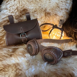 Puede incluir: Un bolso de cuero marrón con cierre de hebilla, unas gafas de cuero marrón con lentes transparentes y una bombilla de luz de cuerda sobre un fondo de piel sintética marrón.