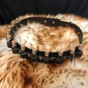 Puede incluir: Un cinturón de cuero marrón con una fila de pequeños viales de vidrio con tapones de corcho. Los viales se mantienen en su lugar mediante remaches metálicos. El cinturón está sobre una superficie peluda marrón y blanca.