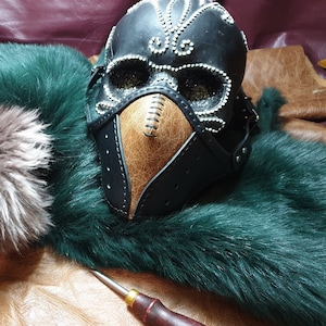 May include: A black leather mask with a brown leather beak and silver studs. The mask is sitting on a green fur background.