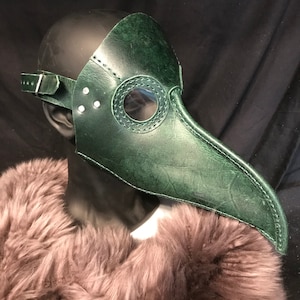 May include: A green leather plague doctor mask with a long beak and a round eye hole. The mask is on a mannequin head.