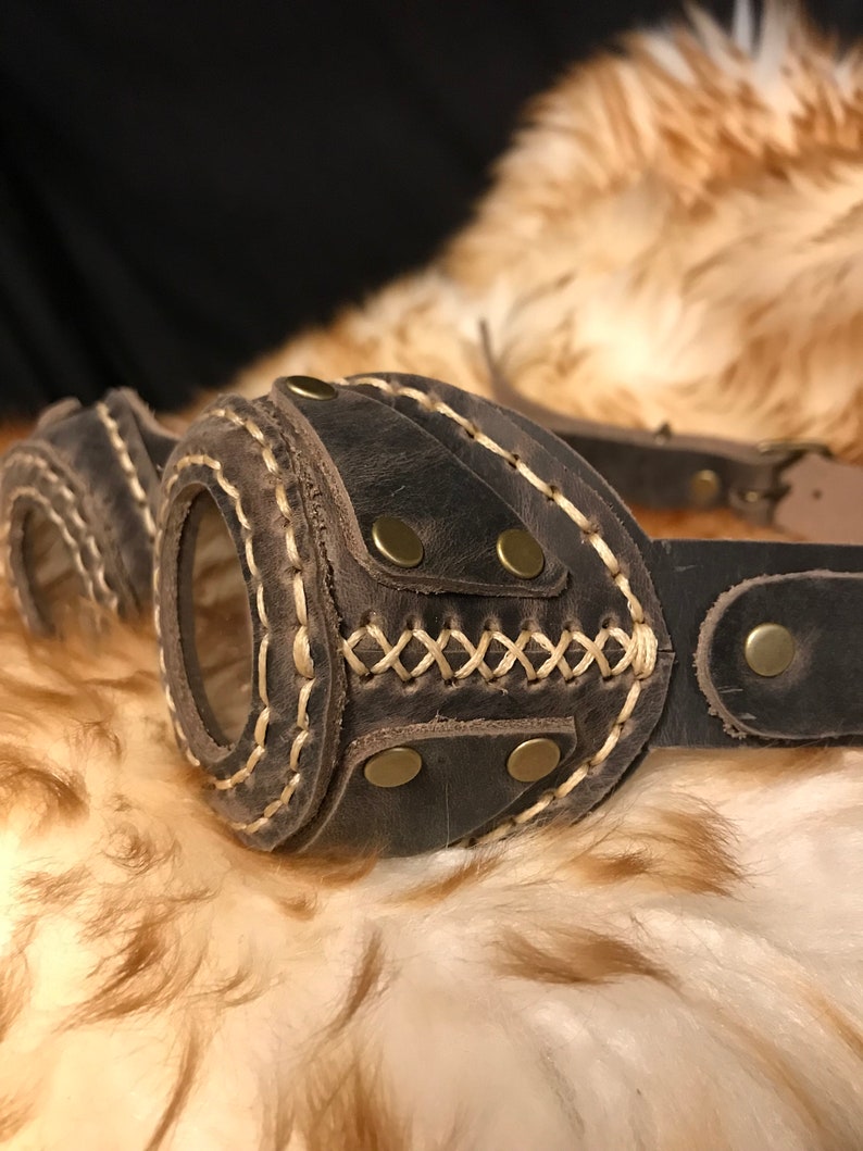 May include: Brown leather steampunk goggles with gold rivets and stitching. The goggles have a round lens and are attached to a leather strap.