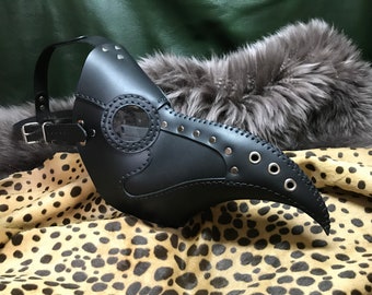 May include: A black leather plague doctor mask with a clear plastic eye piece, metal rivets, and eyelets. The mask has a long beak and is secured with a strap.