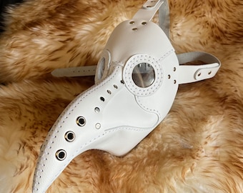 May include: White leather plague doctor mask with a long beak and a clear plastic eye shield. The mask has multiple eyelets and rivets around the beak and eye shield.