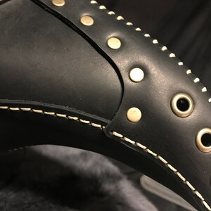 May include: Close-up of a black leather mask with white stitching and gold-toned rivets. The mask has a long, pointed nose and eye holes.
