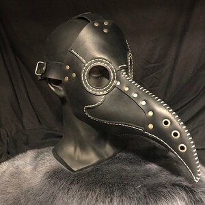 May include: A black leather plague doctor mask with a long beak, white stitching, and gold-toned rivets. The mask has a clear eye piece and a strap for the head.