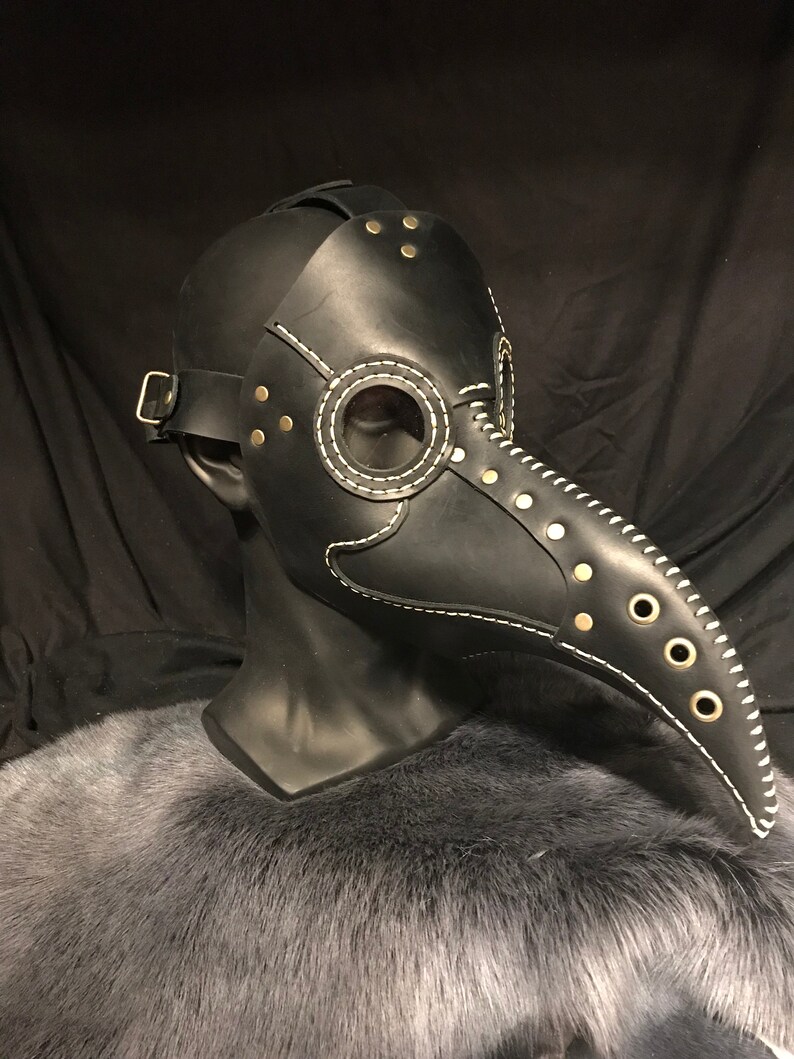 May include: A black leather plague doctor mask with a long beak, white stitching, and gold-toned rivets. The mask has a clear eye piece and a strap for the head.
