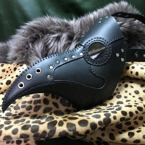 Puede incluir: Una máscara de médico de la peste de cuero negro con un pico largo, agujeros para los ojos y un protector ocular de plástico transparente. La máscara tiene ojales y remaches de metal y está decorada con costuras.