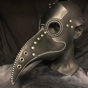 May include: A black leather plague doctor mask with a long beak, white stitching, and gold-toned rivets. The mask is on a black mannequin head.