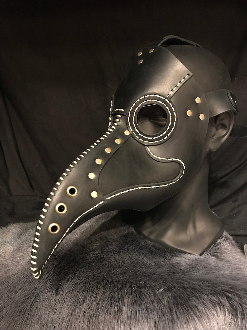 May include: A black leather plague doctor mask with a long beak, white stitching, and gold-toned rivets. The mask is on a black mannequin head.