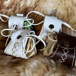 May include: A selection of small leather pouches in white, brown, and gold, with white stitching and drawstring closures. The pouches have a button closure and are displayed on a textured, brown surface.