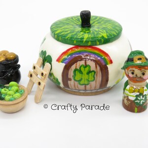 May include: A wooden leprechaun figurine with a green hat, holding a pot of gold. The figurine is standing in front of a wooden pot with a green shamrock design and a rainbow painted on the side. The pot has a lid with a green shamrock design. There is a small wooden ladder and a bowl of green beads next to the pot.
