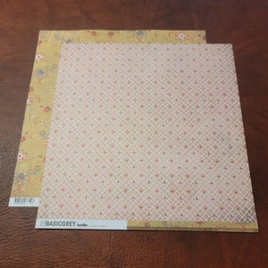 May include: Two sheets of patterned scrapbook paper. One sheet has a floral border in yellow and a cream background with a grid pattern. The other sheet has a cream background with a grid pattern. The brand name "BASICGREY" is visible.