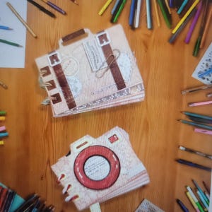 May include: Two handmade journals shaped like a suitcase and a camera, resting on a wooden surface. The suitcase journal is tan with brown accents and a handle. The camera journal has a red lens and a floral pattern. Pencils and other art supplies surround the journals.