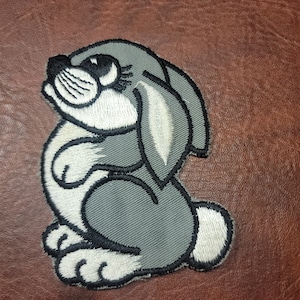 May include: A gray embroidered patch of a cartoon rabbit with white fur and black eyes. The rabbit is sitting with its paws tucked in front of it.