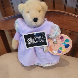 May include: A plush teddy bear wearing a lavender dress with floral sleeves and holding a wooden artist's palette with colorful paint. The bear has a tag that reads "Muffy VanderBear".