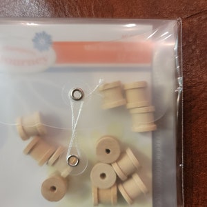 May include: A clear plastic package containing several small, unfinished wooden spools. The spools are cylindrical with a hole through the center. The package is sealed with a string and metal eyelets. The background is a brown leather-like surface.