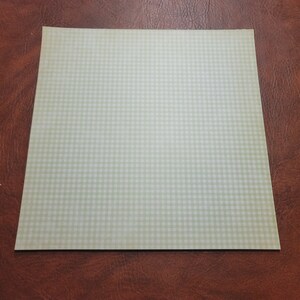 May include: A square sheet of light green and white gingham patterned paper. The paper is lying on a brown leather surface. The gingham pattern is a classic design, often used for crafts and scrapbooking.