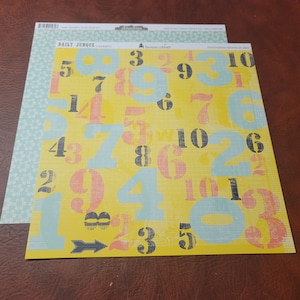 May include: Two sheets of patterned scrapbook paper. One sheet has a yellow background with large numbers in black, blue, and peach. The other sheet has a light blue background with a floral pattern. The text "DAILY JUNQUE" is visible.