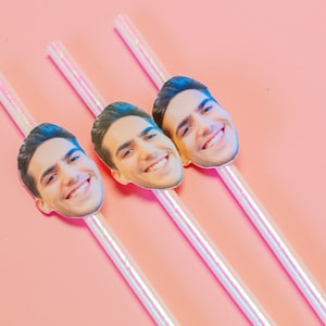 May include: Three iridescent drinking straws with a photo of a person's face on each straw. The faces are smiling and the straws are on a pink background.