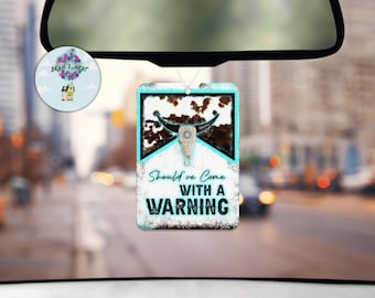 May include: A white car air freshener with a brown and white cow skull design. The text on the air freshener reads "Should've Come With A Warning".
