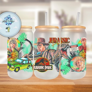 May include: Frosted glass beverage cups with bamboo lids and stainless steel straws. Each cup features a Jurassic Park design with characters, a jeep, and the movie logo. The cups are decorated with vibrant colors and tropical foliage.
