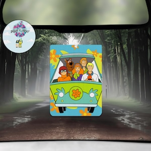 May include: A green and blue car air freshener with a cartoon image of the Scooby Doo gang riding in the Mystery Machine. The characters are Fred, Daphne, Velma, Shaggy, and Scooby Doo.