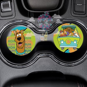 May include: Two round car coasters featuring a cartoon image of Scooby Doo and the Mystery Machine. The Scooby Doo coaster has a green, yellow, and orange background with the words "Ruh Roh" repeated. The Mystery Machine coaster has a green background with the Mystery Machine driving through a field of flowers.