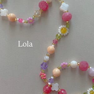 May include: A beaded necklace featuring a variety of colorful elements including pink, peach, white, and lavender round beads, clear crystal accents, and small white and yellow daisy-shaped beads. The word Lola is printed in white text on the left.