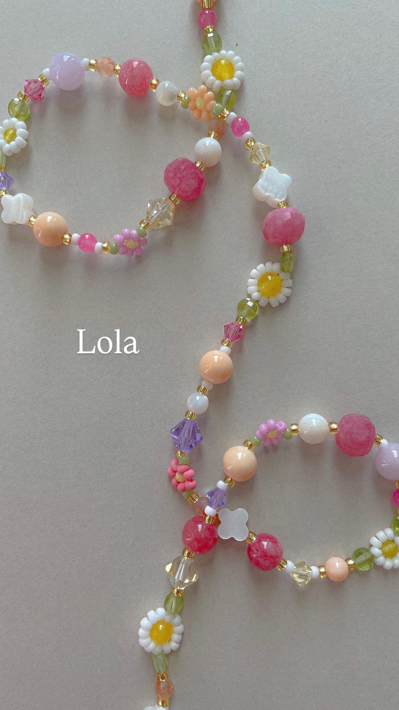 May include: A beaded necklace featuring a variety of colorful elements including pink, peach, white, and lavender round beads, clear crystal accents, and small white and yellow daisy-shaped beads. The word Lola is printed in white text on the left.